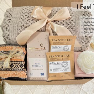 May include: A gift basket filled with cozy items, including a throw blanket, a pair of socks, tea, chocolate, and a candle. The basket is decorated with a beige ribbon and the text "I Feel You".