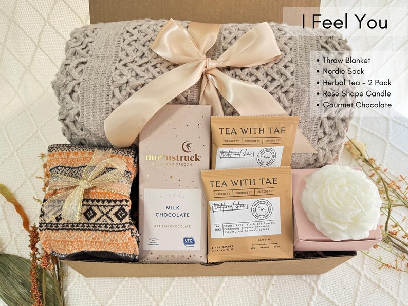 May include: A gift basket filled with cozy items, including a throw blanket, a pair of socks, tea, chocolate, and a candle. The basket is decorated with a beige ribbon and the text "I Feel You".
