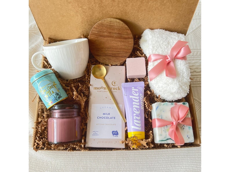 May include: A gift box filled with self-care items including a white mug, a tin of Earl Grey tea, a pink candle, a bar of milk chocolate, a lavender hand cream, a pink bath bomb, a white fluffy sock with a pink ribbon, and a bar of soap with a pink ribbon.