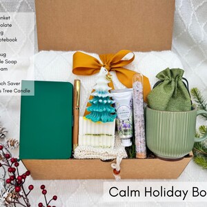 Calm Holiday Box featuring notebook, pen, white blanket, green mug, Christmas tree candle, bath salt tube, hand cream, handmade soap, and burlap bag styled on white winter background