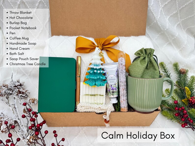 Calm Holiday Box featuring notebook, pen, white blanket, green mug, Christmas tree candle, bath salt tube, hand cream, handmade soap, and burlap bag styled on white winter background