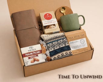 May include: A gift box containing a brown leather-style journal, a box of Lark Salted Caramel Almond Chocolate Chip cookies, a green ribbed mug with a gold spoon, a pair of patterned knit socks, a bag of Tea With Tae, and Young's Praline Pecans.