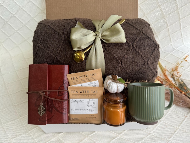 Peut inclure: Une couverture en tricot marron, deux bo&icirc;tes de th&eacute; &eacute;tiquet&eacute;es "Tea with Tae", un journal en cuir marron avec un charme en forme de feuille, une tasse en c&eacute;ramique verte, un petit bocal en verre avec une bougie et une citrouille d&eacute;corative blanche.