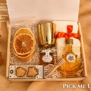 May include: A gift box containing a gold candle holder, white fluffy socks with an orange ribbon, a bag of dried orange slices, a jar of honey, a wooden honey stirrer, a maple leaf-shaped bottle of maple syrup, and a box of maple leaf candies.