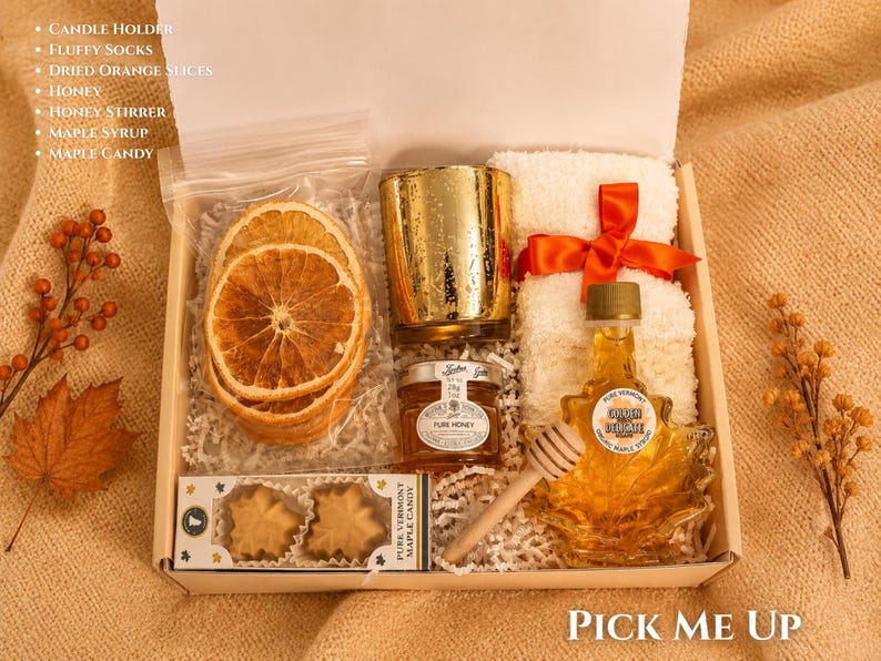May include: A gift box containing a gold candle holder, white fluffy socks with an orange ribbon, a bag of dried orange slices, a jar of honey, a wooden honey stirrer, a maple leaf-shaped bottle of maple syrup, and a box of maple leaf candies.