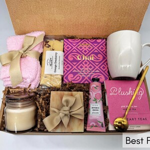 May include: A gift box filled with self-care items, including a pink washcloth, a bar of orange cranberry soap, a pink hand cream tube, a pink tea bag box, a white mug, a candle, and a bar of soap. The box is decorated with brown paper shreds and a gold ribbon.