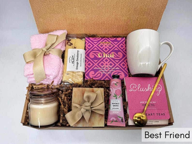 May include: A gift box filled with self-care items, including a pink washcloth, a bar of orange cranberry soap, a pink hand cream tube, a pink tea bag box, a white mug, a candle, and a bar of soap. The box is decorated with brown paper shreds and a gold ribbon.