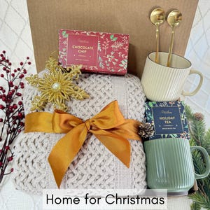 May include: A gift basket with a cozy beige knitted throw blanket, gold snowflake ornament, two gold spoons, a white mug, a green mug, and two boxes of tea and chocolate. The boxes have the text "Chocolate Chip" and "Holiday Tea" on them. The basket is labeled "Home for Christmas".