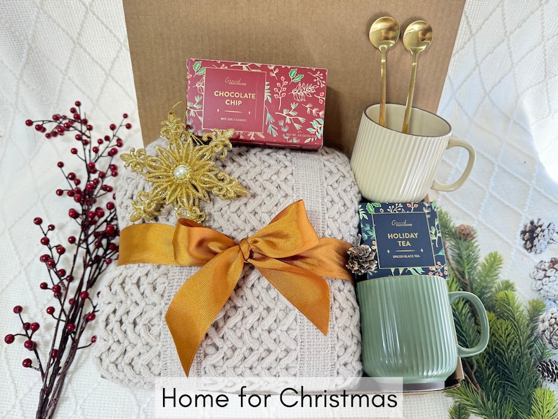 May include: A gift basket with a cozy beige knitted throw blanket, gold snowflake ornament, two gold spoons, a white mug, a green mug, and two boxes of tea and chocolate. The boxes have the text "Chocolate Chip" and "Holiday Tea" on them. The basket is labeled "Home for Christmas".