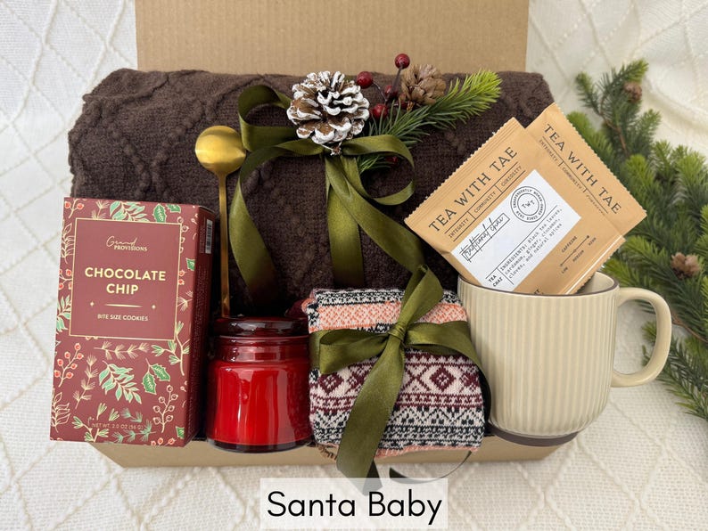 May include: A gift basket with a brown knitted blanket, a red candle, a box of chocolate chip cookies, a mug, and two packages of tea labeled "Tea with Tae".