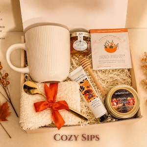 May include: A gift box containing a white ribbed coffee mug, a jar of honey, a wooden honey stirrer, a packet of Lavender Orange Grey herbal tea, a tube of shea butter hand cream, a gold spoon, and fluffy socks tied with a red ribbon.