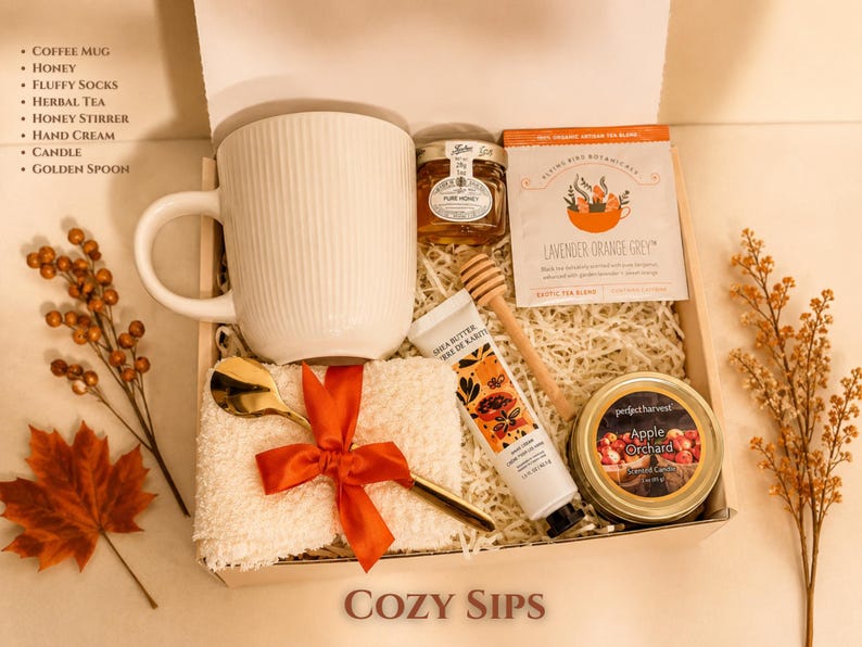 May include: A gift box containing a white ribbed coffee mug, a jar of honey, a wooden honey stirrer, a packet of Lavender Orange Grey herbal tea, a tube of shea butter hand cream, a gold spoon, and fluffy socks tied with a red ribbon.