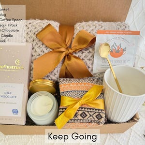 May include: A gift box filled with self-care items, including a throw blanket, coffee mug, tea, chocolate, candle, and socks. The box is tied with a gold ribbon and the words "Keep Going" are printed on the front.