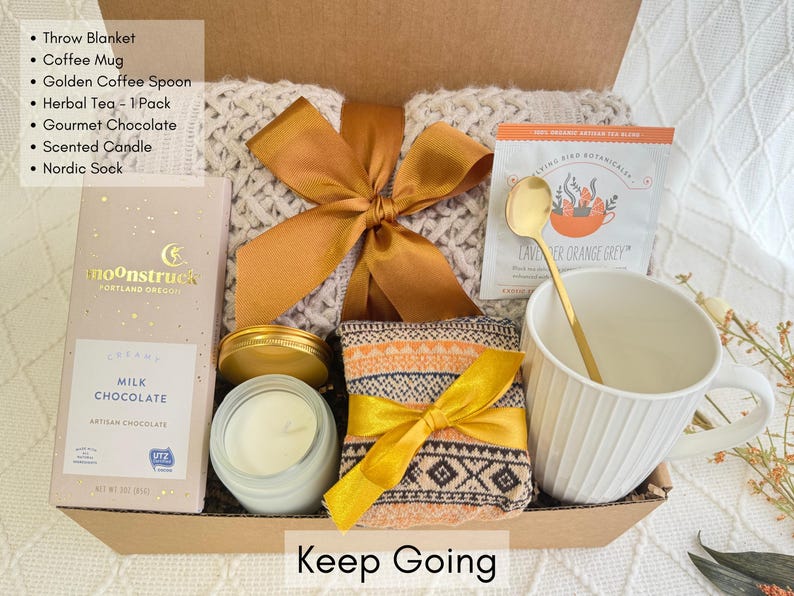 May include: A gift box filled with self-care items, including a throw blanket, coffee mug, tea, chocolate, candle, and socks. The box is tied with a gold ribbon and the words "Keep Going" are printed on the front.