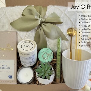 May include: A white gift box filled with self-care items, including a throw blanket, a white mug, a candle, a bath bomb, a succulent, a bar of chocolate, a tea bag, and bath salts. The box is decorated with a green ribbon and a tag that says "Joy Gift Box".