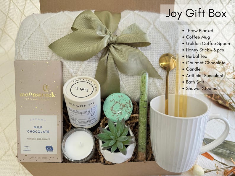 May include: A white gift box filled with self-care items, including a throw blanket, a white mug, a candle, a bath bomb, a succulent, a bar of chocolate, a tea bag, and bath salts. The box is decorated with a green ribbon and a tag that says "Joy Gift Box".