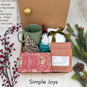 Puede incluir: Una caja de regalo navide&ntilde;a con una taza verde, galletas con chispas de chocolate, t&eacute;, un adorno navide&ntilde;o, una cuchara dorada, una vela y calcetines blancos y esponjosos. Decorada con bayas rojas y ramas de pino.
