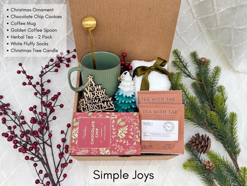 Puede incluir: Una caja de regalo navide&ntilde;a con una taza verde, galletas con chispas de chocolate, t&eacute;, un adorno navide&ntilde;o, una cuchara dorada, una vela y calcetines blancos y esponjosos. Decorada con bayas rojas y ramas de pino.