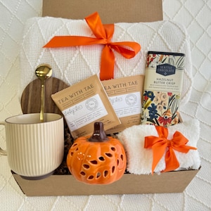 Peut inclure: Un panier-cadeau rempli d'articles sur le th&egrave;me de l'automne, comprenant une couverture blanche, une tasse, un bougeoir en forme de citrouille, deux sachets de th&eacute;, une tablette de chocolat et une serviette blanche avec un ruban orange.