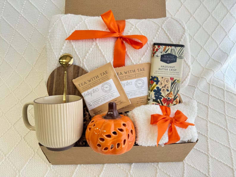 Peut inclure: Un panier-cadeau rempli d'articles sur le th&egrave;me de l'automne, comprenant une couverture blanche, une tasse, un bougeoir en forme de citrouille, deux sachets de th&eacute;, une tablette de chocolat et une serviette blanche avec un ruban orange.