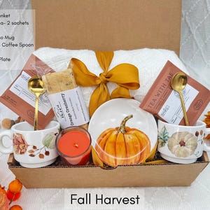 K&ouml;nnte beinhalten: Eine herbstliche Geschenkbox mit einer wei&szlig;en Decke, Kr&auml;utertee, Biscotti, Espressotassen, goldenen L&ouml;ffeln, einer Kerze und einem Mini-Teller. Der Teller zeigt ein K&uuml;rbismotiv, und die Box ist mit einem goldenen Band gebunden. Der Text "Fall Harvest" steht unten.