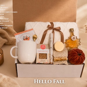 May include: A gift box containing a white ribbed mug, a cream throw blanket with a brown ribbon, a bottle of maple syrup, chocolate chip cookies, a soap saver, herbal tea, handmade soap, a shower steamer, bath salt, a pumpkin, and a golden spoon.