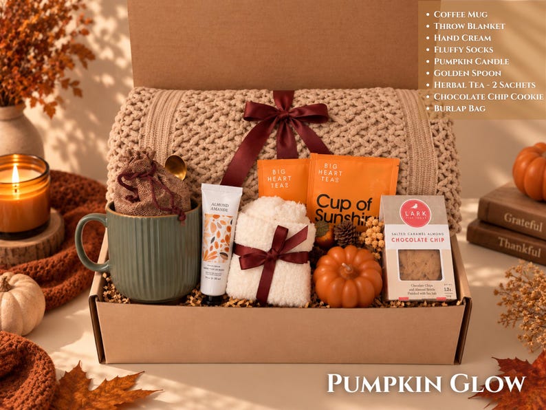 May include: A gift box containing a beige knit throw blanket with a maroon ribbon, a green ribbed mug, a burlap bag, a tube of hand cream, two orange tea sachets, white fluffy socks, a small orange pumpkin, and a box of chocolate chip cookies.