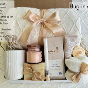 May include: A white gift box filled with self-care items, including a throw blanket, a mug, a candle, soap, chocolate, hand cream, and a fluffy sock. The box is labeled "Hug in a Box".