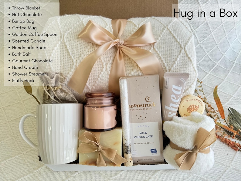 May include: A white gift box filled with self-care items, including a throw blanket, a mug, a candle, soap, chocolate, hand cream, and a fluffy sock. The box is labeled "Hug in a Box".