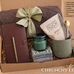 May include: A gift box containing a brown textured throw blanket with a green ribbon, a dark brown journal with a gold pen, a green candle, patterned socks, a green ribbed coffee mug, a gold spoon, and a bag of Wolfgang Puck Sorrento coffee.