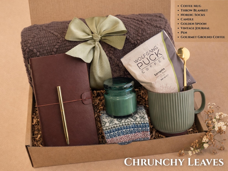 May include: A gift box containing a brown textured throw blanket with a green ribbon, a dark brown journal with a gold pen, a green candle, patterned socks, a green ribbed coffee mug, a gold spoon, and a bag of Wolfgang Puck Sorrento coffee.