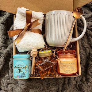 May include: A gift box filled with self-care items, including a white mug, a small tin of tea, a honey jar, a candle, a wooden honey dipper, and a white towel with a brown ribbon.