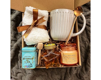 May include: A gift box filled with self-care items, including a white mug, a small tin of tea, a honey jar, a candle, a wooden honey dipper, and a white towel with a brown ribbon.