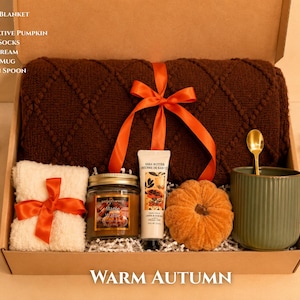 May include: A gift box labeled WARM AUTUMN containing a brown diamond-patterned throw blanket tied with an orange ribbon, a jar candle, a tube of hand cream, a decorative orange pumpkin, fluffy white socks, a green mug, and a golden spoon.