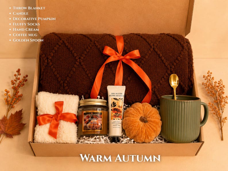 May include: A gift box labeled WARM AUTUMN containing a brown diamond-patterned throw blanket tied with an orange ribbon, a jar candle, a tube of hand cream, a decorative orange pumpkin, fluffy white socks, a green mug, and a golden spoon.