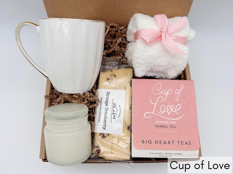 May include: A gift box filled with a white mug, a candle, a package of orange cranberry biscotti, a white hand towel with a pink ribbon, and a pink box of "Cup of Love" herbal tea.