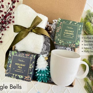 Puede incluir: Una caja de regalo con una taza blanca, una toalla blanca atada con una cinta verde y dos cajas de golosinas. Tambi&eacute;n se incluye una vela de &aacute;rbol de Navidad verde y blanca y bayas rojas. El texto "Jingle Bells" est&aacute; en la parte inferior.
