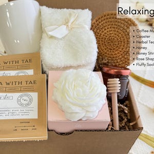 May include: A gift box filled with relaxing items, including a white coffee mug, two tea sachets labeled "TEA WITH TAE", a white fluffy sock, a white rose-shaped candle, a small jar of honey with a wooden honey dipper, a white fluffy towel, and a woven coaster.