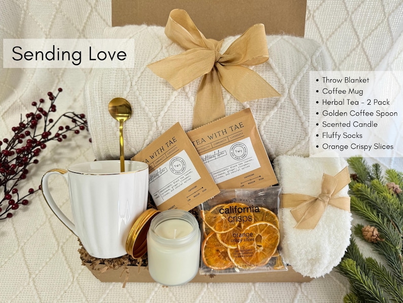 May include: A white gift box filled with a white throw blanket, a white coffee mug, two tea packets, a golden coffee spoon, a white candle, a pair of white fluffy socks, and a bag of orange slices. The box is tied with a brown ribbon and the text "Sending Love" is written on the top of the box.