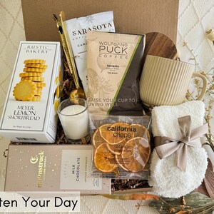 May include: A gift basket filled with various items, including a mug, a candle, shortbread cookies, chocolate, orange slices, and a small white towel with a brown ribbon. The basket is decorated with brown paper and wood.