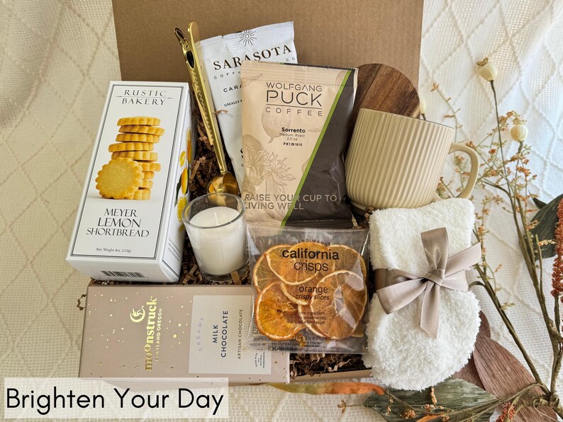 May include: A gift basket filled with various items, including a mug, a candle, shortbread cookies, chocolate, orange slices, and a small white towel with a brown ribbon. The basket is decorated with brown paper and wood.
