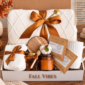May include: A gift box labeled FALL VIBES containing a white textured throw blanket with a brown ribbon, a white ribbed coffee mug, two tea sachets, a jar of honey with a wooden stirrer, a small candle, a wooden coaster, and fluffy white socks.