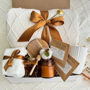 Peut inclure: Un panier-cadeau blanc rempli d'articles confortables pour une soir&eacute;e relaxante. Il comprend une couverture blanche, une serviette blanche, une tasse, une bougie, un pot de miel, un sous-verre en bois et deux sachets de th&eacute;. Les sachets de th&eacute; sont &eacute;tiquet&eacute;s "Tea with Tae".