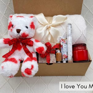 May include: A gift basket filled with a red and white striped teddy bear, a white blanket, a candle, bath salts, and a bar of soap. The basket is decorated with a red ribbon and a white bow. The text "I love you more" is visible at the bottom of the image.