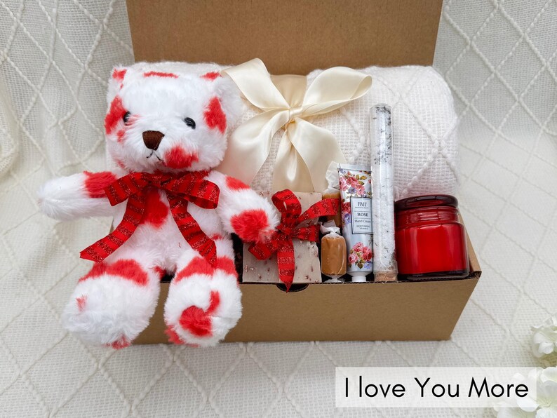 May include: A gift basket filled with a red and white striped teddy bear, a white blanket, a candle, bath salts, and a bar of soap. The basket is decorated with a red ribbon and a white bow. The text "I love you more" is visible at the bottom of the image.