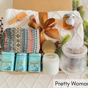 May include: A gift basket filled with a variety of items, including a white blanket, a pair of knitted socks, a jar of honey, a candle, a tea infuser, and three boxes of tea. The basket is decorated with a brown ribbon and a gold bow.