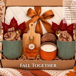 Könnte beinhalten: Eine Geschenkbox mit der Aufschrift Fall Together, die eine cremefarbene Wohndecke mit braunem Band, zwei gerippte salbeigrüne Tassen, ein Holzbrett, eine Ahornblatt-förmige Sirupflasche, eine Kürbisduftkerze, zwei goldene Löffel und zwei Jutesäckchen enthält.