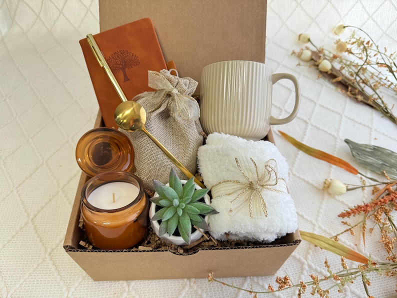 Peut inclure: Une bo&icirc;te-cadeau en carton marron remplie d'une vari&eacute;t&eacute; d'articles, dont un journal en cuir marron, une cuill&egrave;re en or, un petit sac en toile de jute, une tasse en c&eacute;ramique blanche, une serviette blanche, une petite plante succulente en pot et une bougie en verre marron.