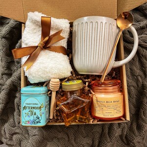 May include: A gift box filled with self-care items, including a white mug, a small tin of tea, a honey jar, a candle, a wooden honey dipper, and a white towel with a brown ribbon.