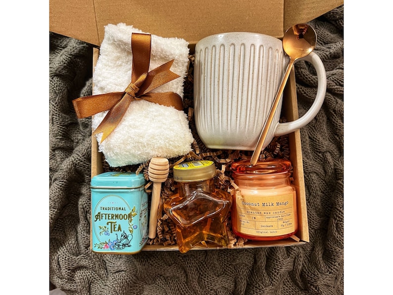 May include: A gift box filled with self-care items, including a white mug, a small tin of tea, a honey jar, a candle, a wooden honey dipper, and a white towel with a brown ribbon.